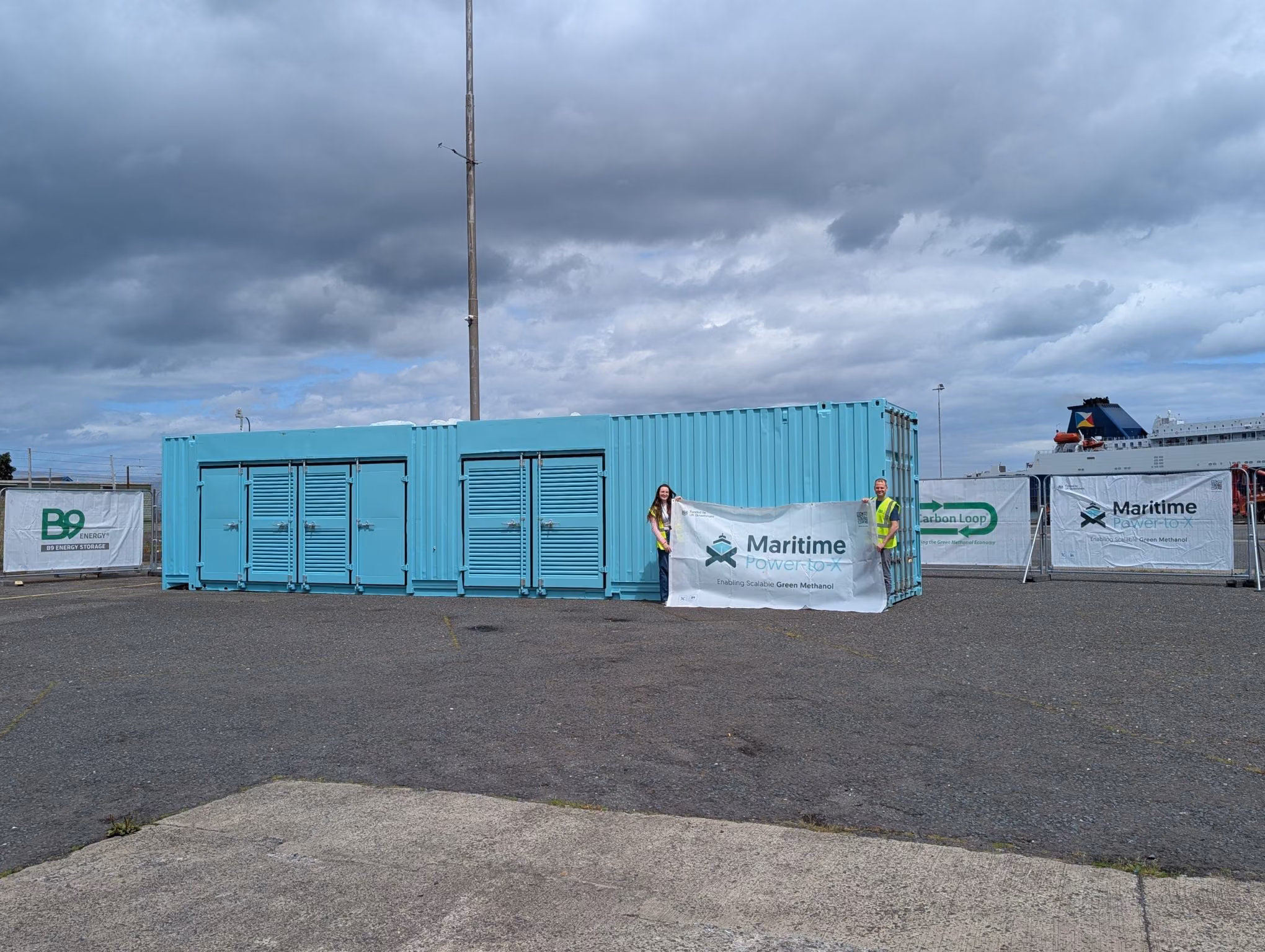 A blue container unit at Larne Harbour with Maritime Power-to-X banners and two people holding a project banner